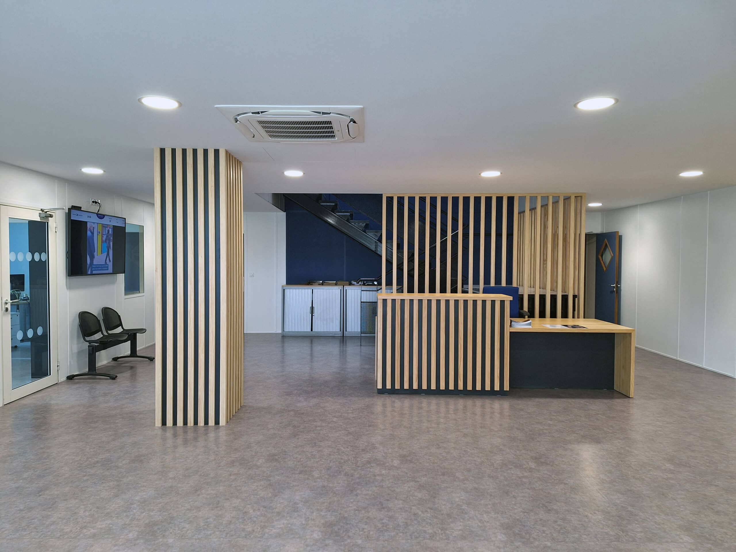 Hall d'accueil moderne avec comptoir en lattes de bois clair, écran affichant 14:50, et escalier bleu visible derrière la structure.