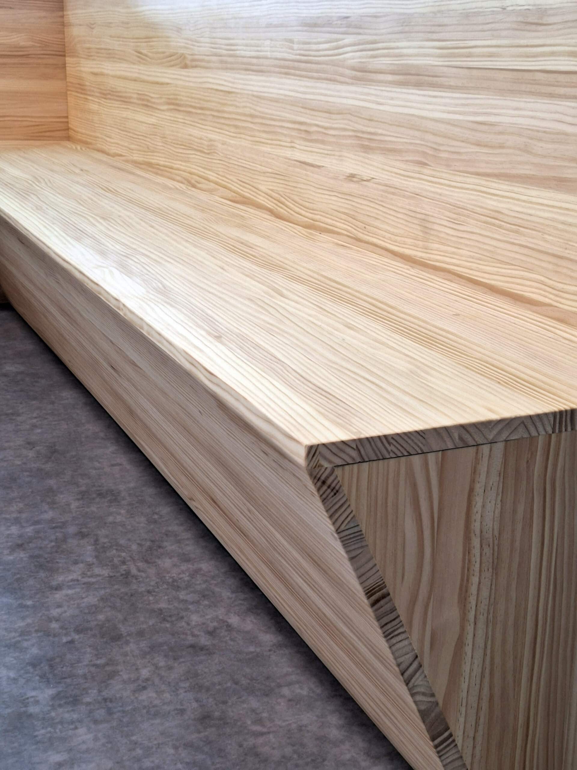 Gros plan d'un banc moderne en bois clair aux veines prononcées, avec une assise et une base inclinée, reposant sur un sol gris foncé.