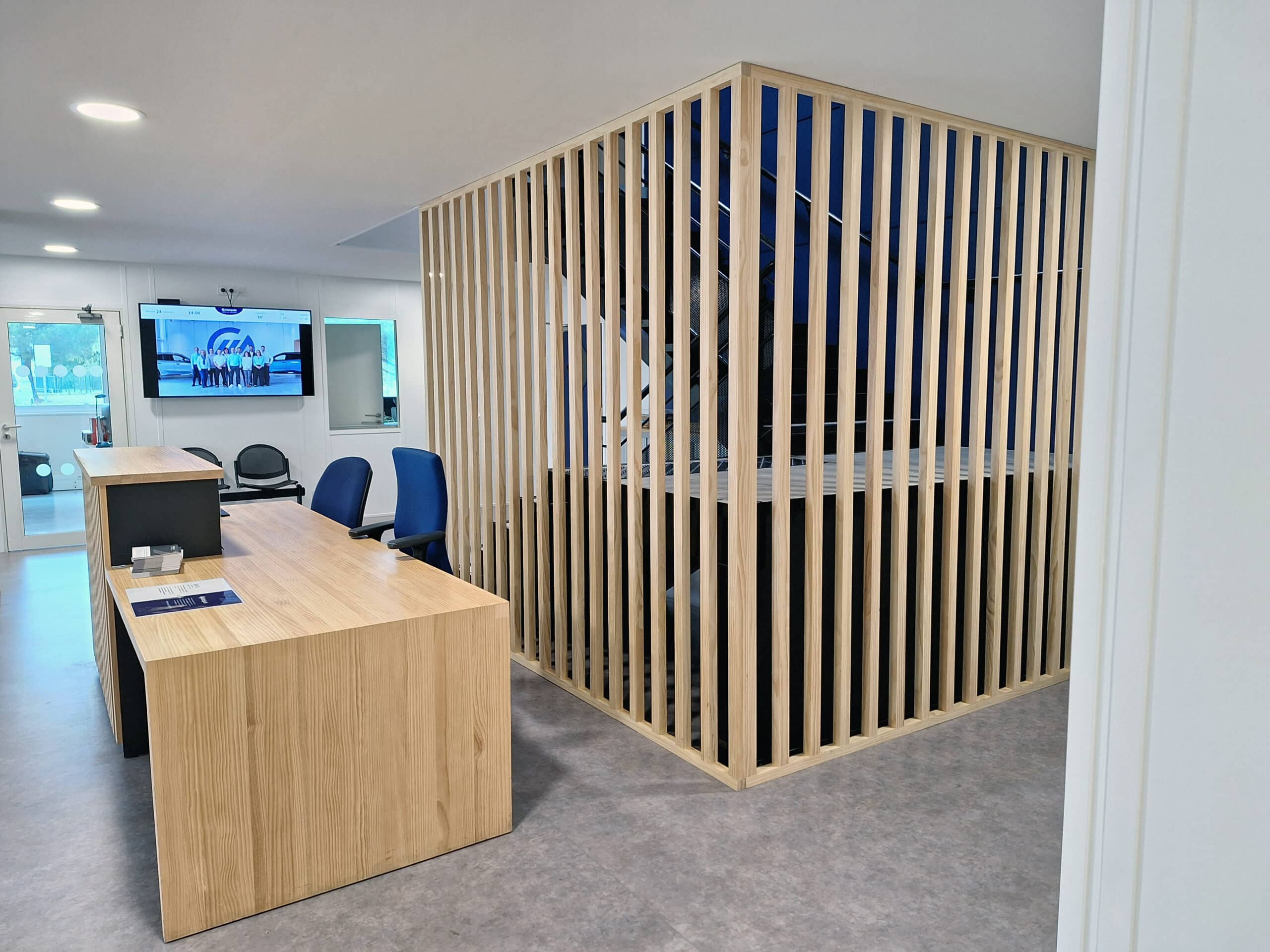 Bureau d'accueil moderne avec comptoir en bois clair, séparateur à claire-voie en bois, chaises bleues, et écran affichant une équipe et la mention "GA".