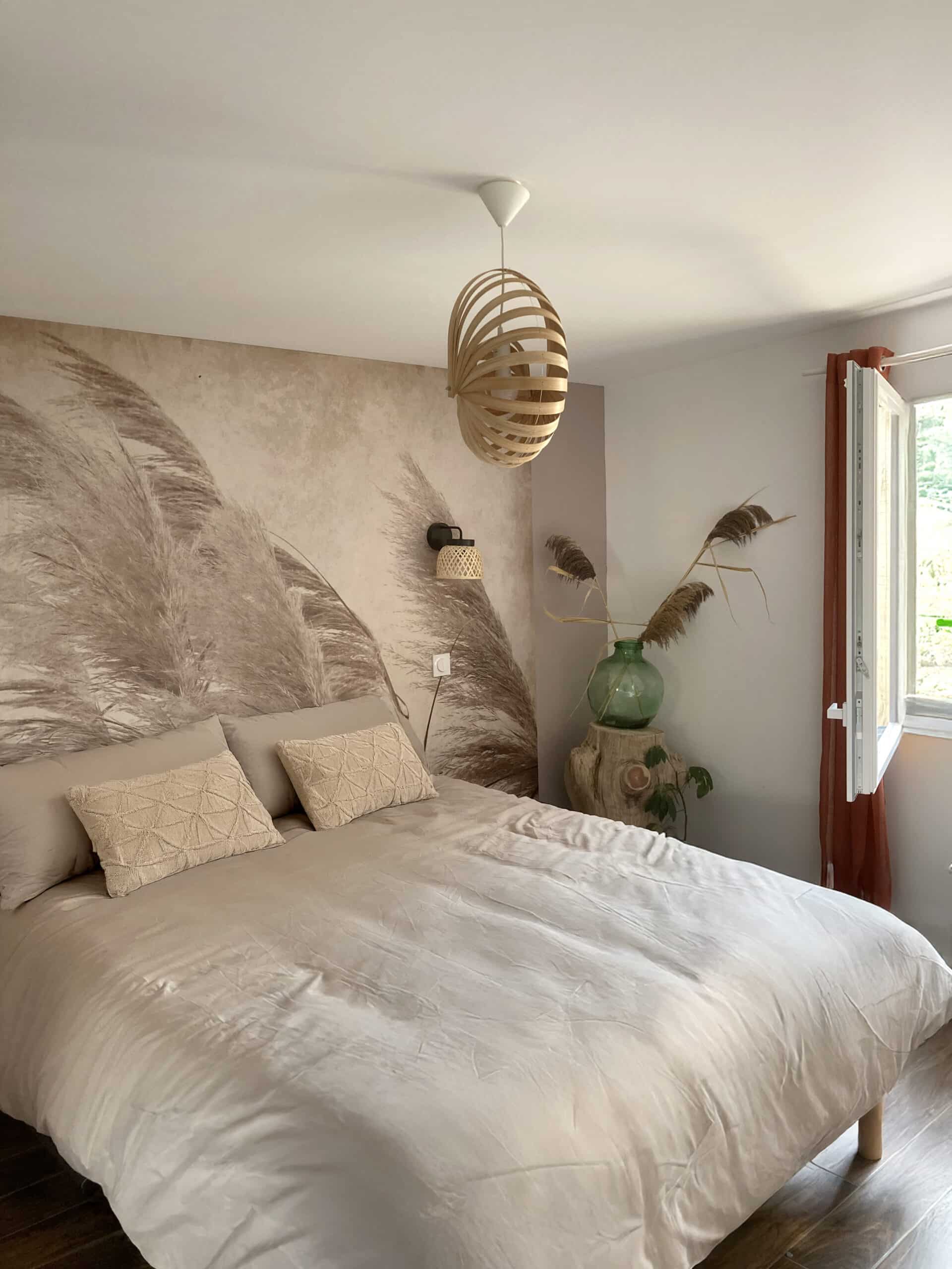 Chambre bohème avec mur papier peint herbe de pampa beige, couette satinée, suspension bois ajouré et fenêtre avec voilage terracotta.