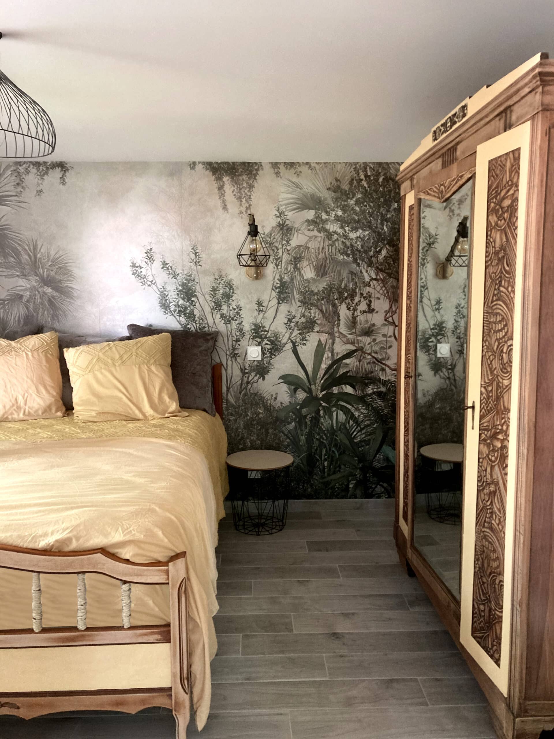 Chambre d'hôtel jungle chic : lit jaune pâle, papier peint panoramique forêt, armoire ancienne sculptée avec miroir et lampe cage.