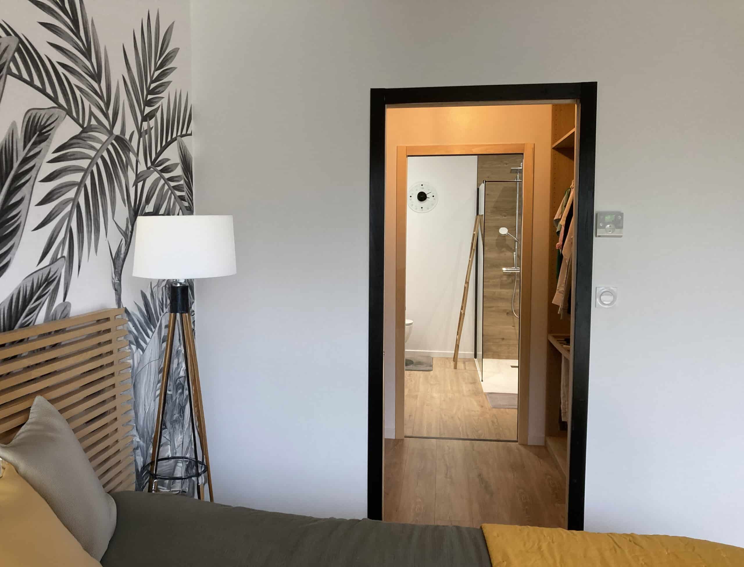 Chambre moderne avec papier peint jungle noir et blanc, lit avec tête de lit en lattes de bois et vue sur une salle de bain/dressing.