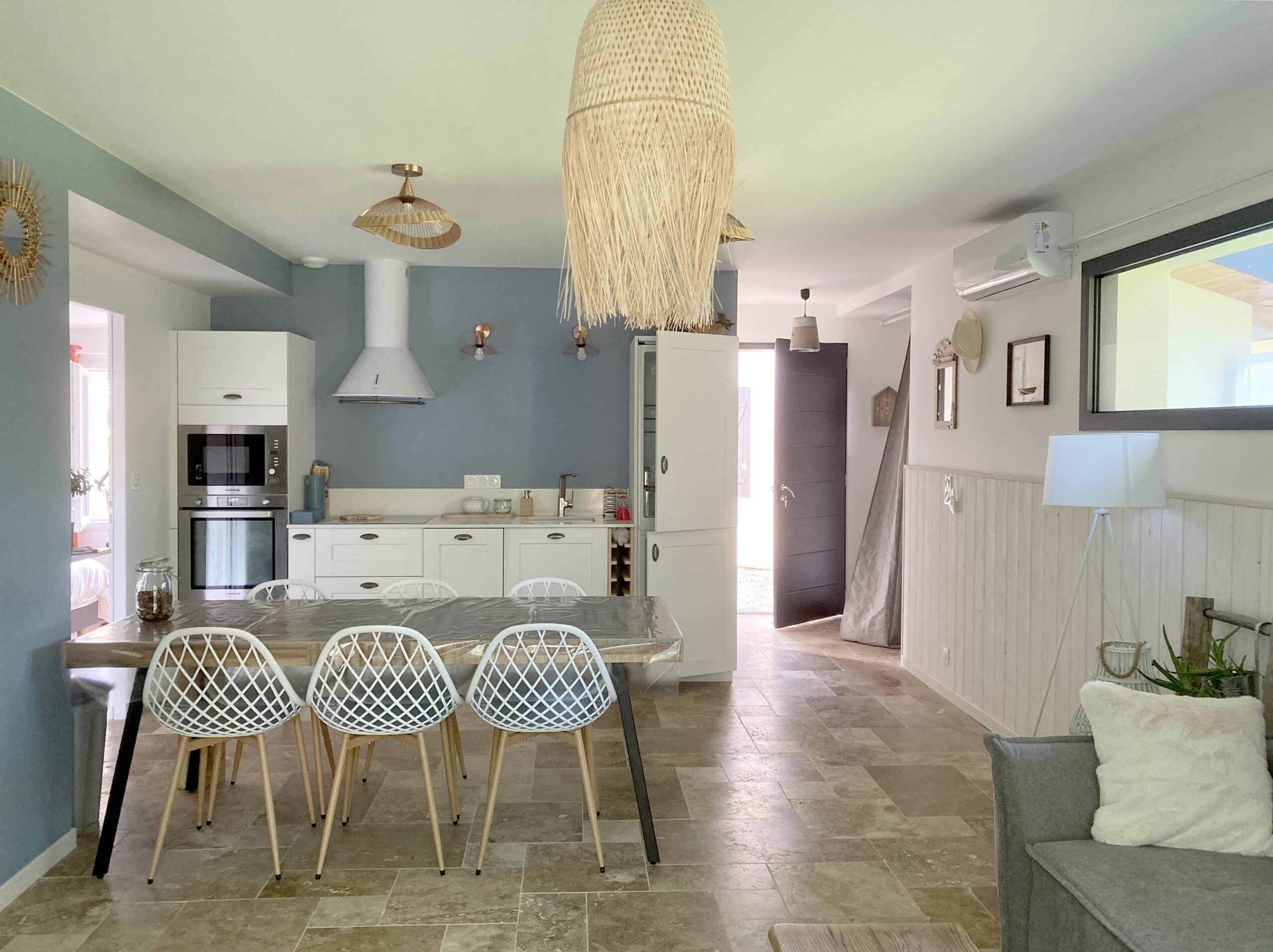 Cuisine moderne ouverte avec murs bleu-gris, mobilier blanc, sol en pierre beige et luminaires en paille naturelle. Chaises de salle à manger blanches.