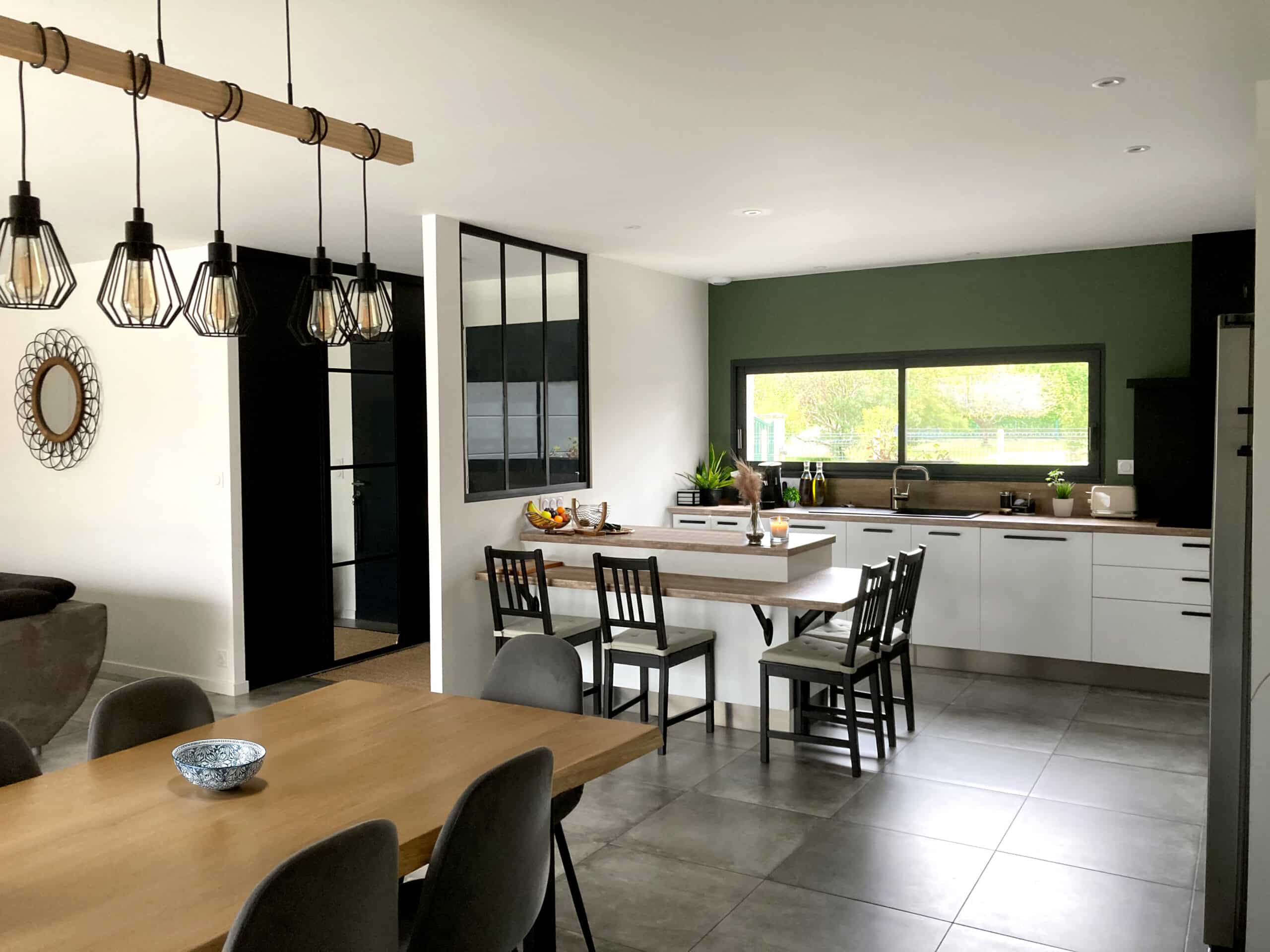 Cuisine moderne ouverte avec murs blancs et accent vert forêt, îlot central bois clair, luminaires noirs style industriel, et table à manger.
