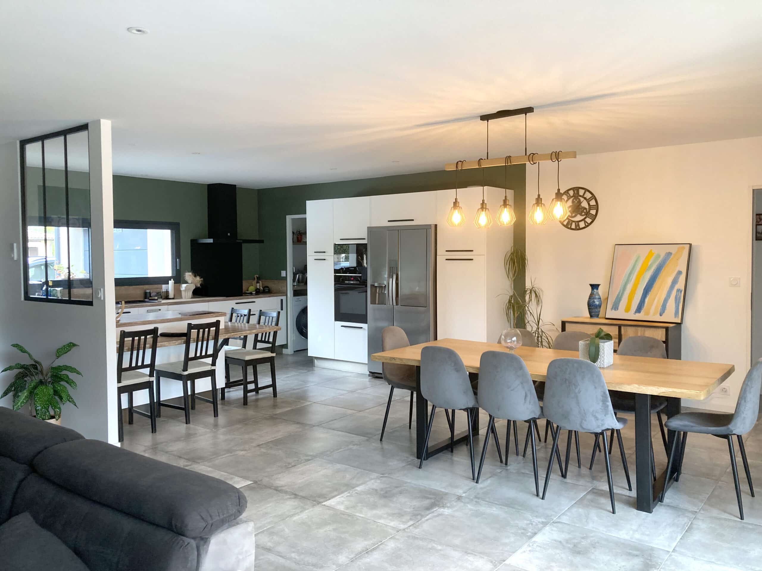 Intérieur moderne avec cuisine ouverte vert sauge, salle à manger contemporaine avec table bois clair, chaises grises et luminaire industriel.