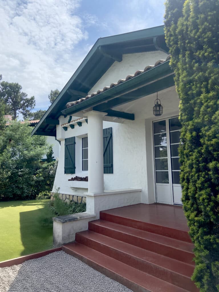 Façade d'une maison basque blanche avec toit en tuiles rouges et poutres vert foncé, près d'un haut buisson taillé.