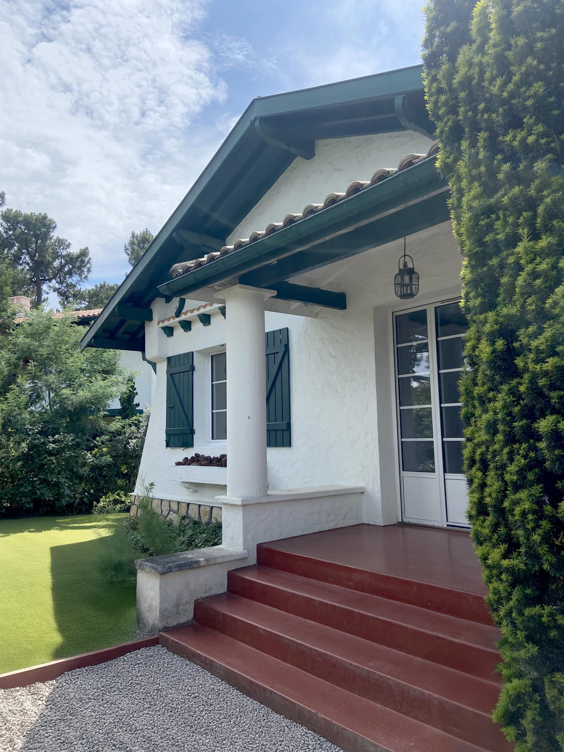 Façade d'une maison basque blanche avec toit en tuiles rouges et poutres vert foncé, près d'un haut buisson taillé.