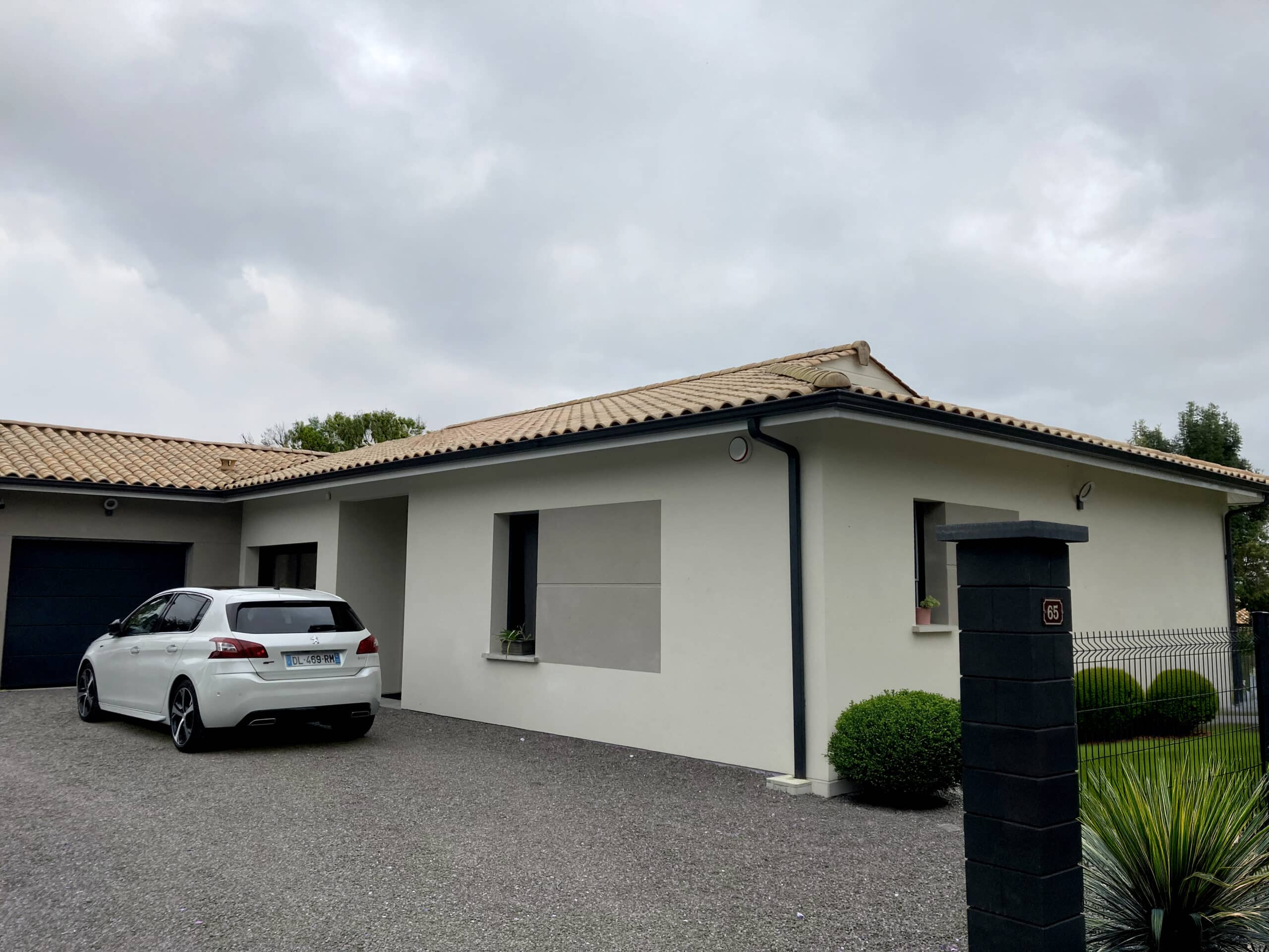 Maison moderne claire avec toit en tuiles romanes, voiture Peugeot blanche garée sur allée gravillonnée et pilier noir portant le numéro 65.
