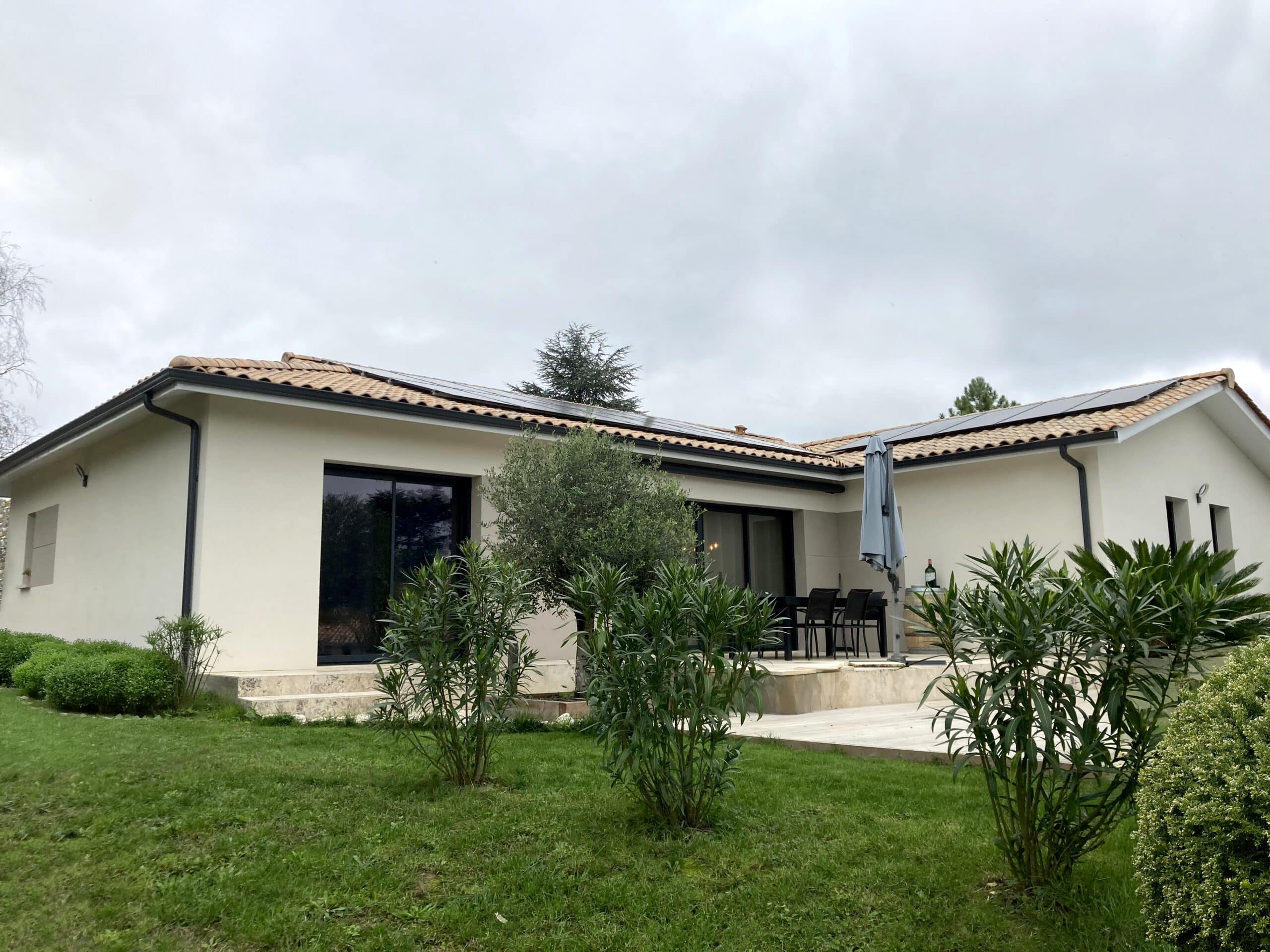 Maison moderne avec façade claire, toit en tuiles romanes et panneaux solaires, vue sur la pelouse verte et la terrasse aménagée.