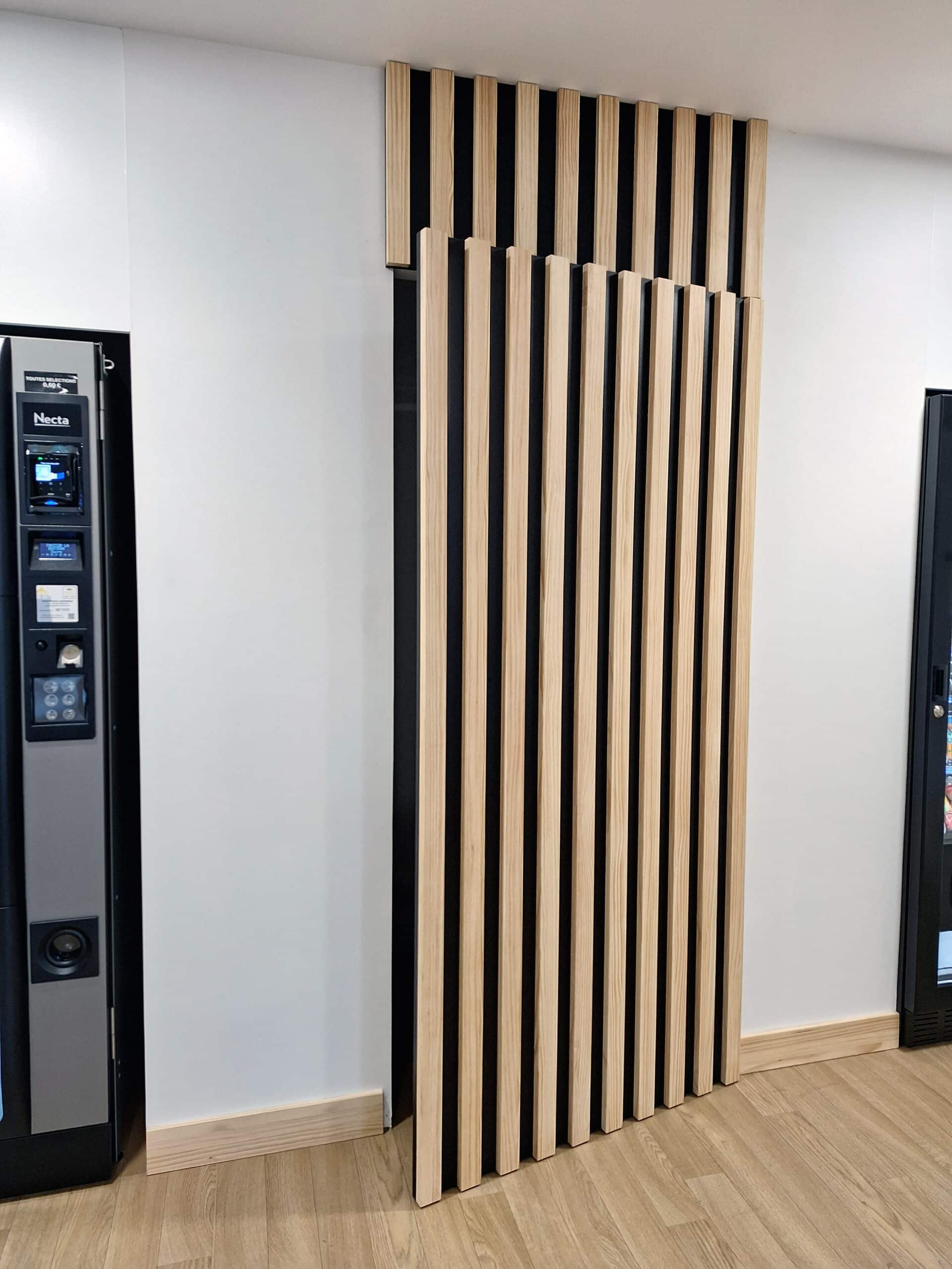 Panneau acoustique à lattes de bois clair sur fond noir, à côté d'un distributeur automatique Necta. Sol stratifié clair.
