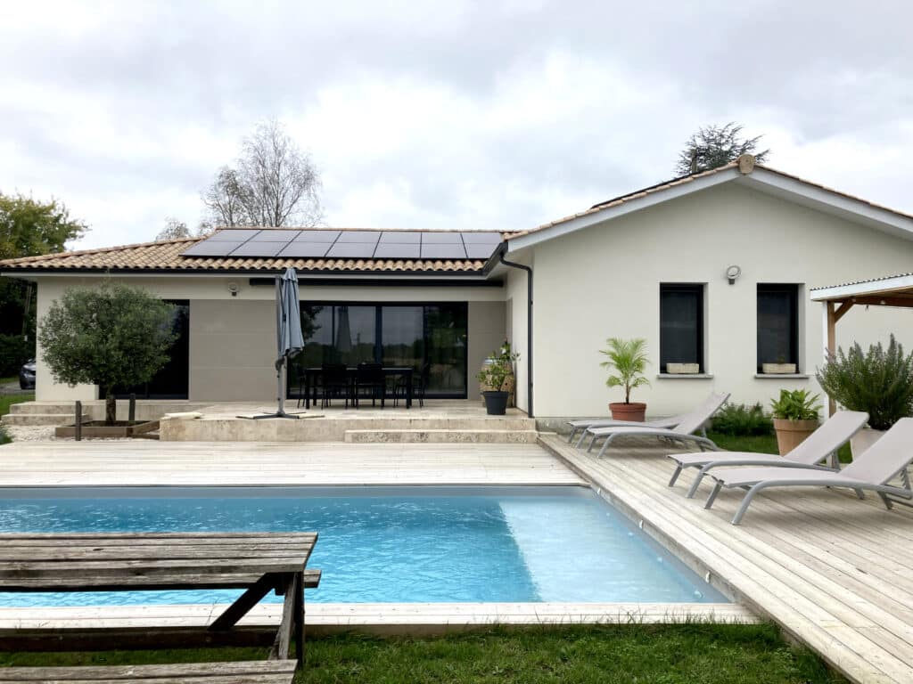 Villa moderne avec piscine, terrasse en bois clair, mobilier de jardin et panneaux solaires sous un ciel nuageux.