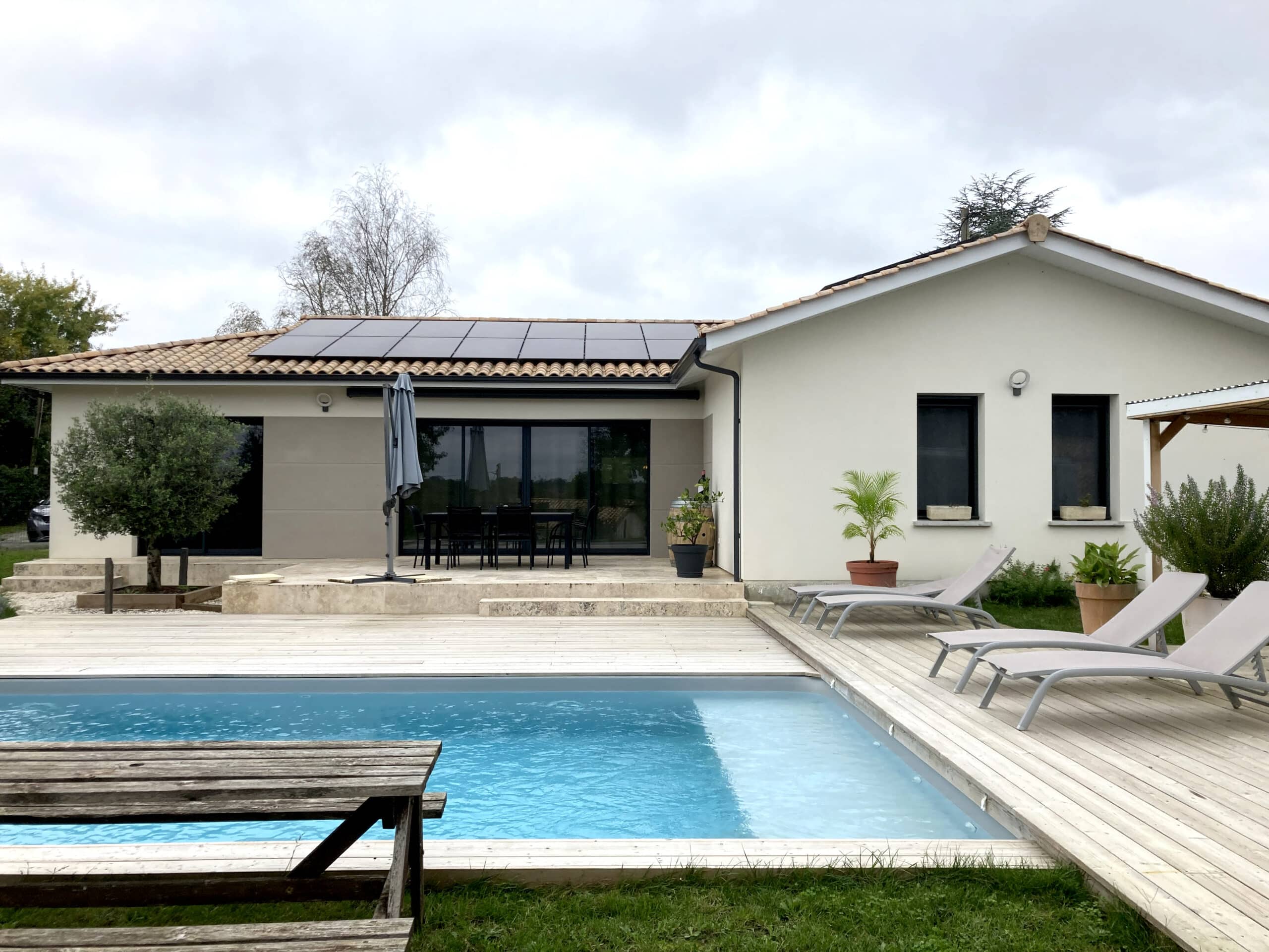 Villa moderne avec piscine, terrasse en bois clair, mobilier de jardin et panneaux solaires sous un ciel nuageux.
