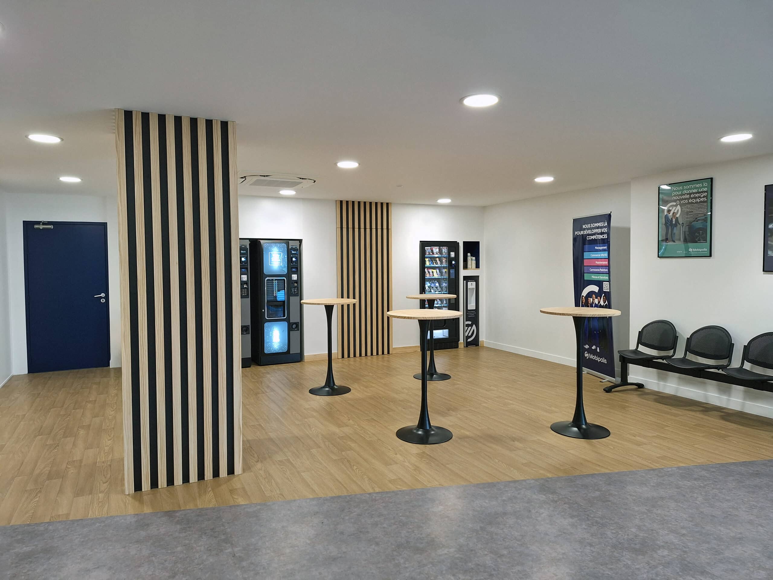 Espace d'attente moderne avec distributeurs automatiques, tables hautes et chaises noires. Panneaux bois/noir et porte bleue.