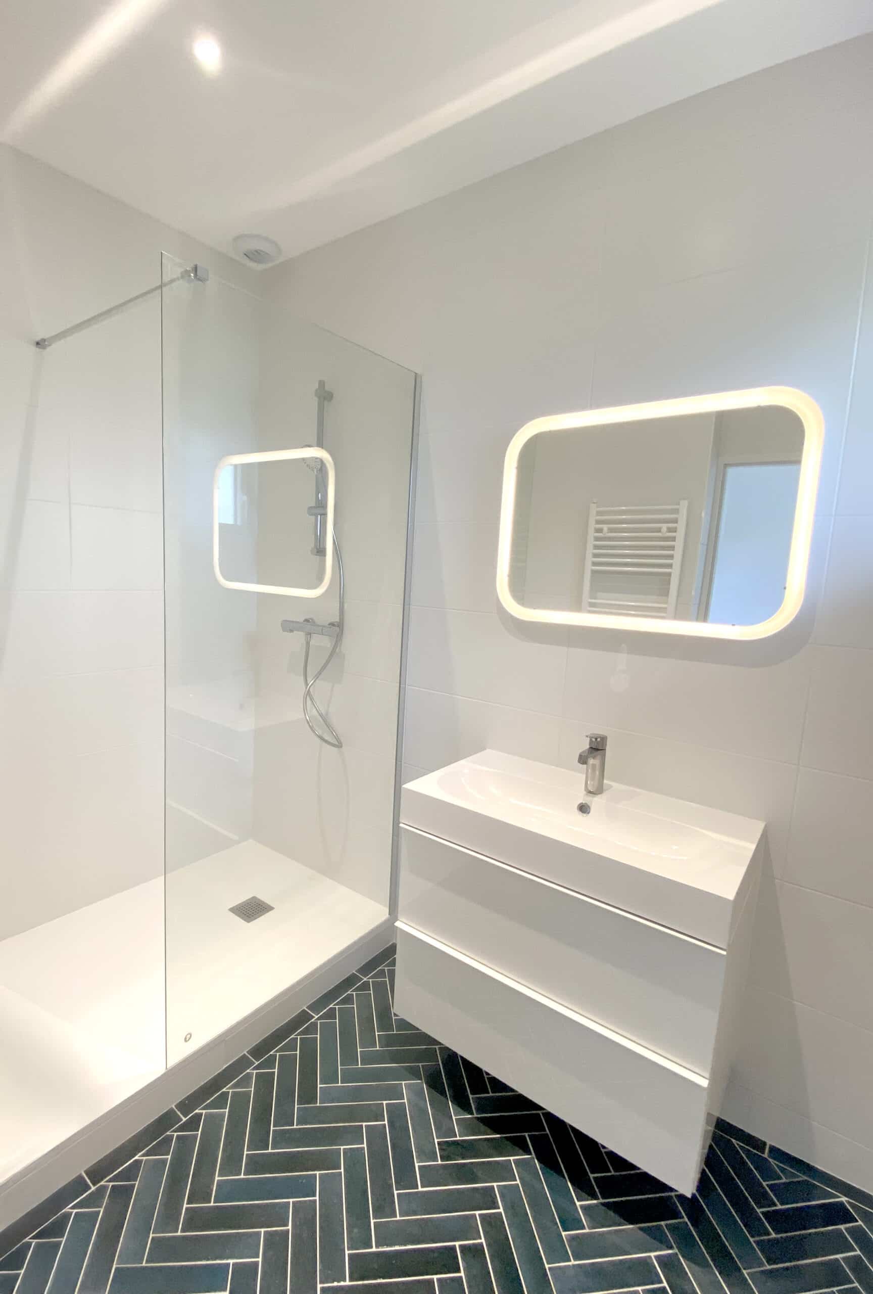 Salle de bain moderne avec douche vitrée, meuble-lavabo blanc, miroir rétroéclairé et sol en carreaux chevrons bleu-vert foncé.
