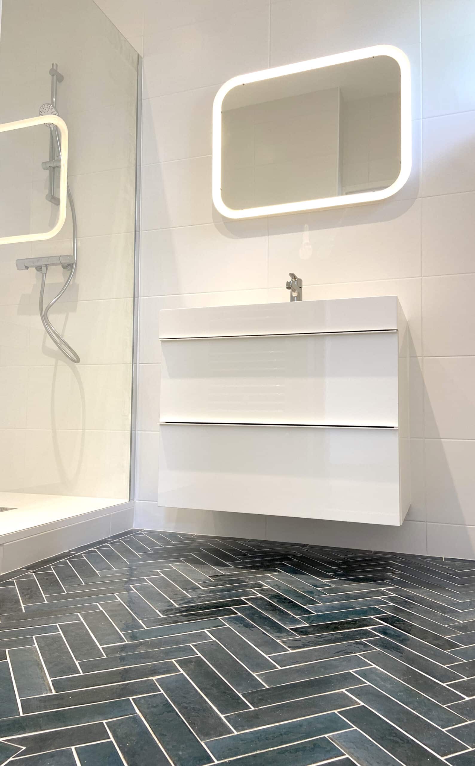 Salle de bain moderne avec meuble-lavabo flottant blanc, miroir lumineux arrondi, et sol en chevrons de carreaux foncés à joints clairs.