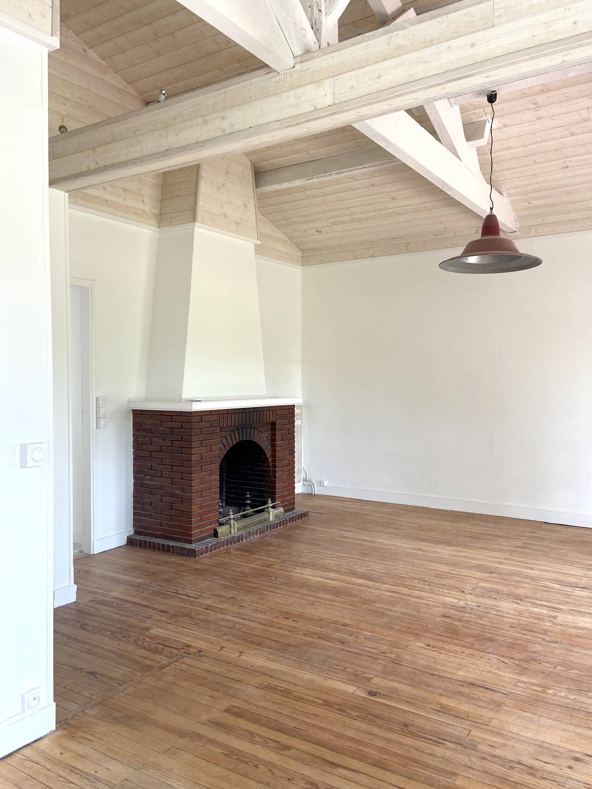 Pièce vide avec parquet bois, murs blancs, grande cheminée en briques rouges et plafond mansardé avec poutres claires et suspension rouge.