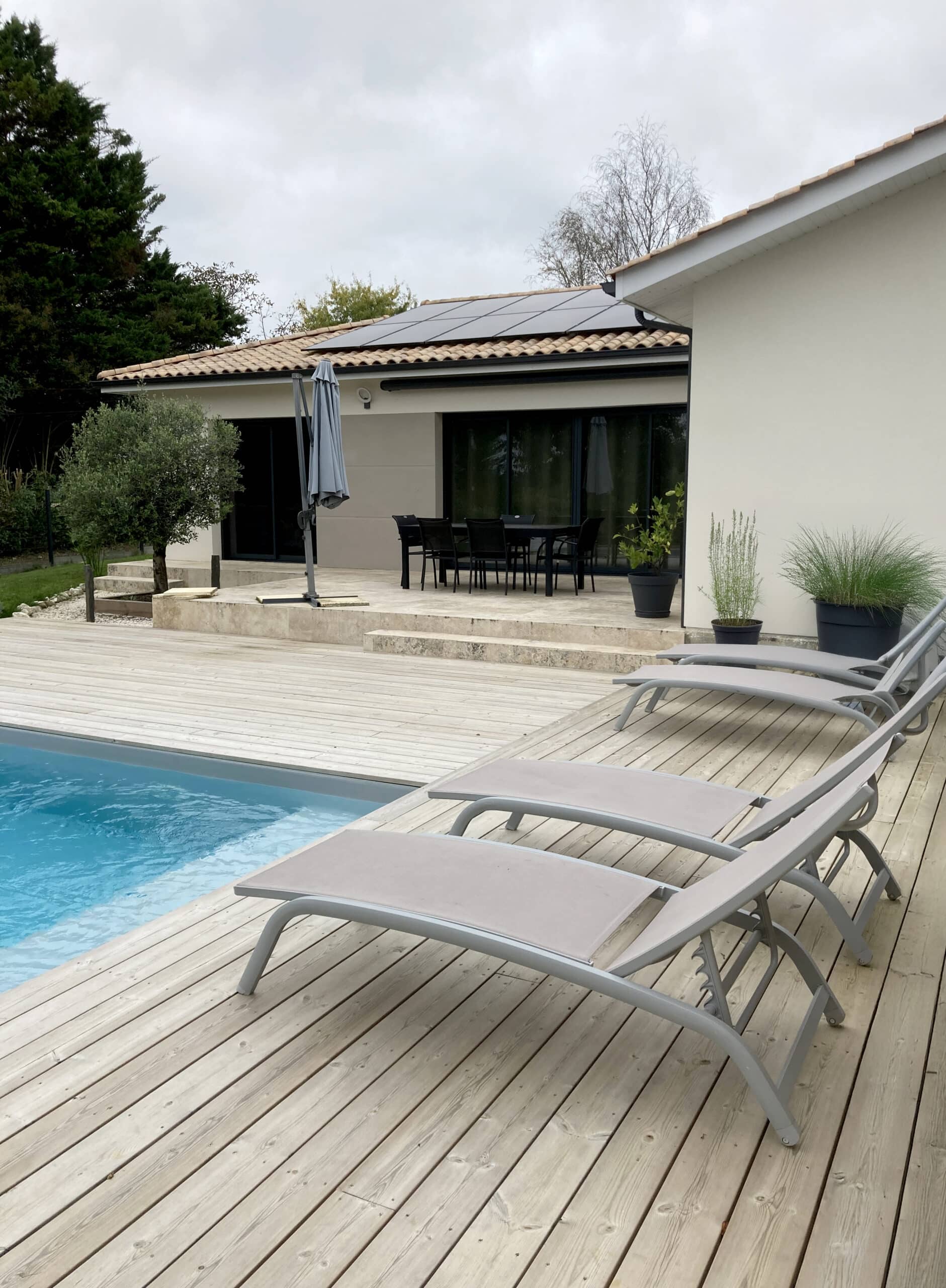 Terrasse en bois clair avec piscine et chaises longues grises, devant une maison moderne aux tuiles rouges et panneaux solaires.